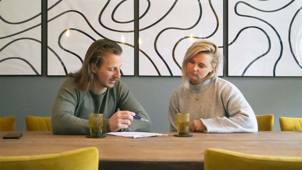 Leefstijl coach die een gesprek voert met een mannelijke client zittend aan een tafel in de woonkamer met schilderijen op de achtergrond.