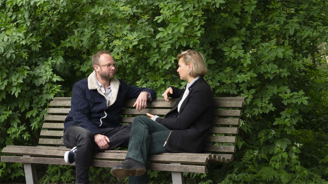 Linde die een gesprek heeft met een client zittend op een bankje met een wand van bladeren op de achtergrond.